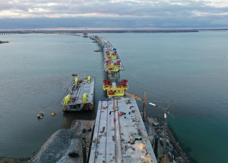 Aerial view of the Dronning Margrethe II bridge construction, with prefabricated bridge segments, construction platforms, and marine works extending across the strait.